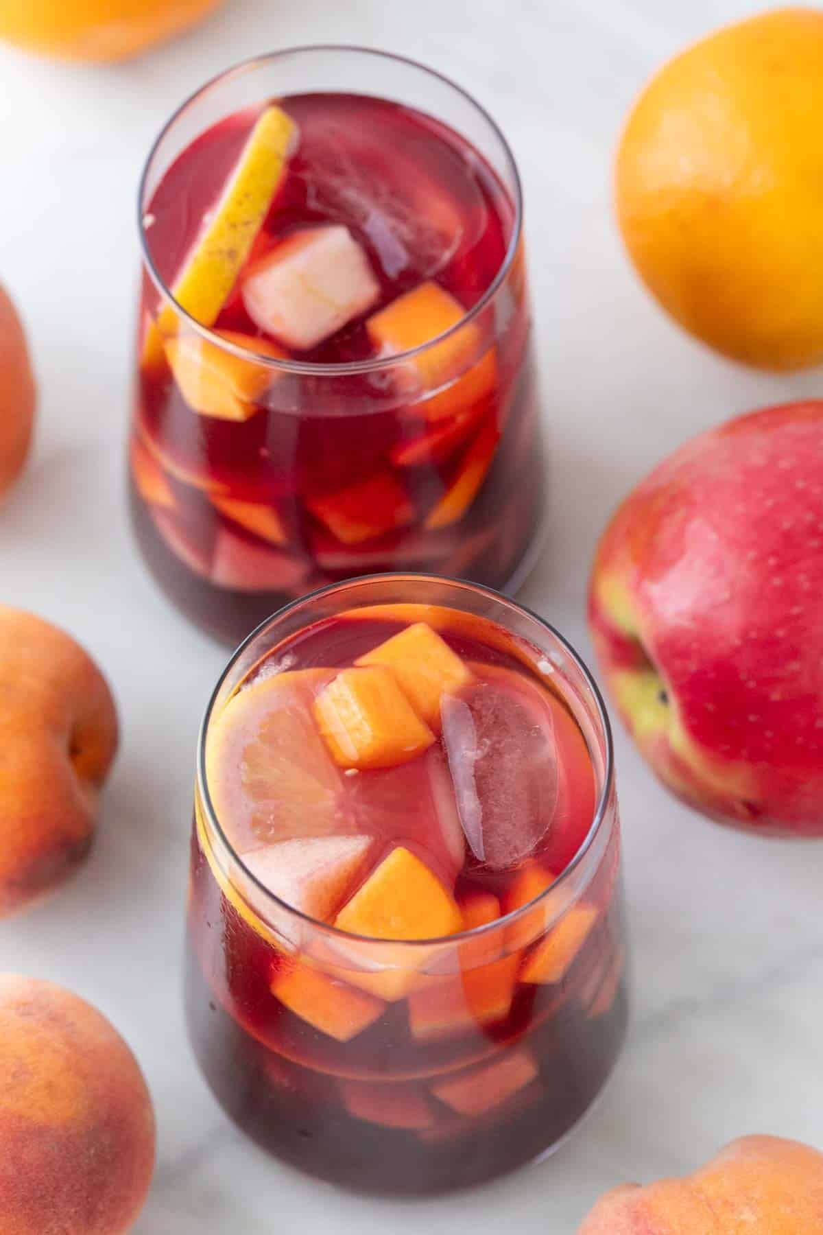 Two glasses of sangria with ice, surrounded by apples, peaches, and oranges
