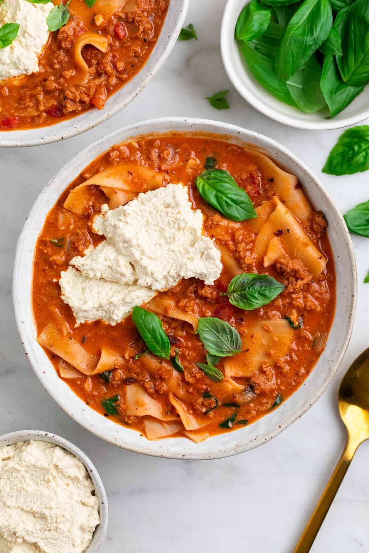 Two bowls of vegan lasagna soup with vegan ricotta, basil, and ingredients around them