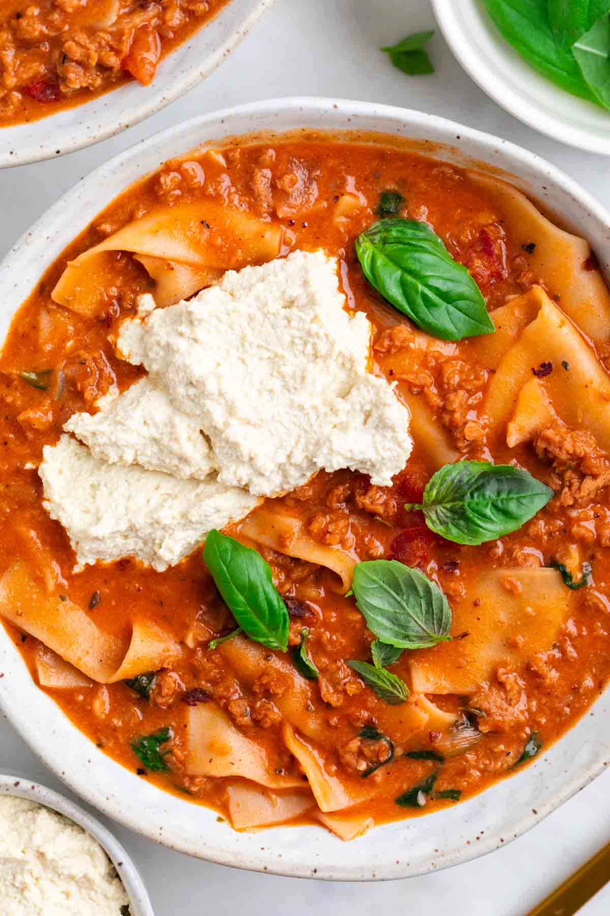 Lasagna soup in two bowls topped with vegan ricotta and basil, with ingredients around
