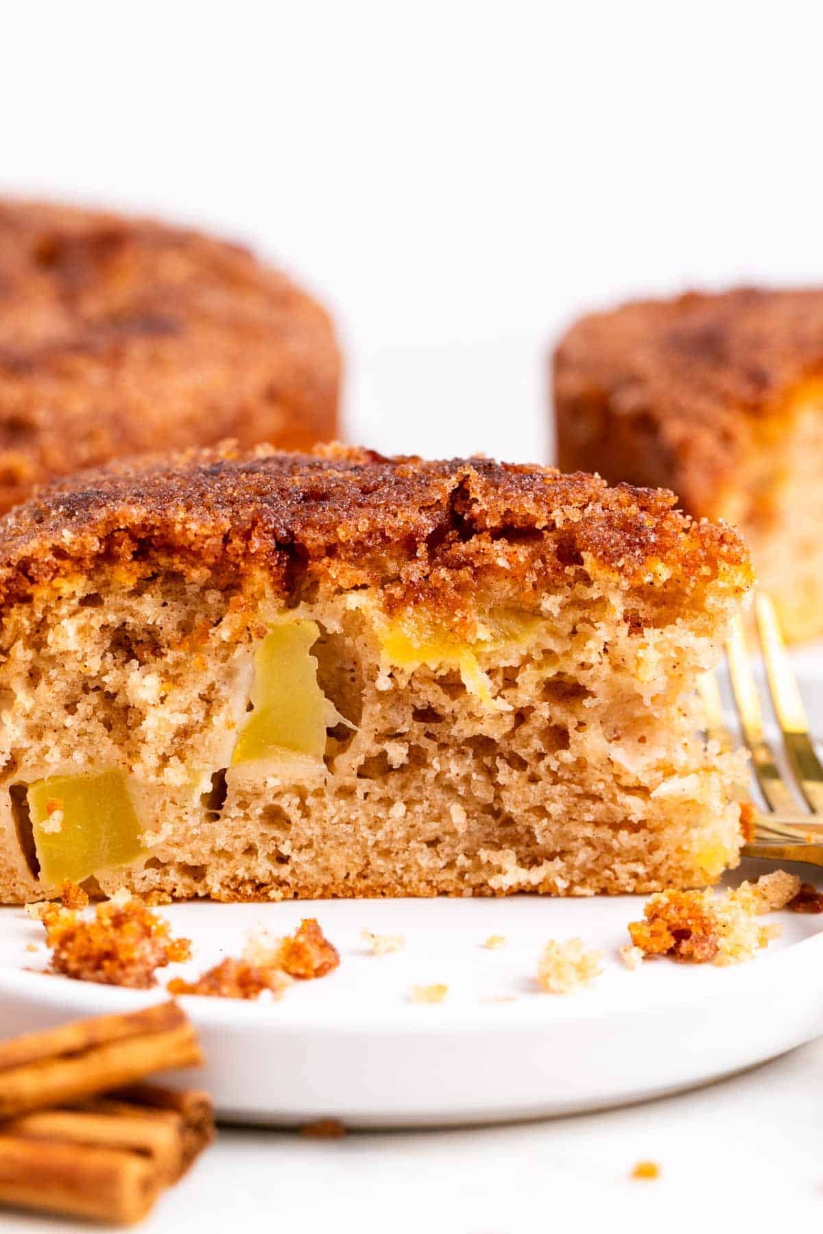 Vegan apple cake slice on a plate with a fork, cinnamon sticks, and cake behind