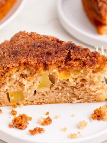Slice of vegan apple cake on a plate with fork and whole cake behind