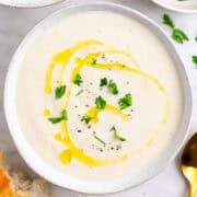 Two bowls of vegan cauliflower soup surrounded by bread, parsley, and a spoon