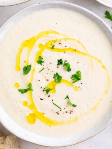 Two bowls of vegan cauliflower soup surrounded by bread, parsley, and a spoon